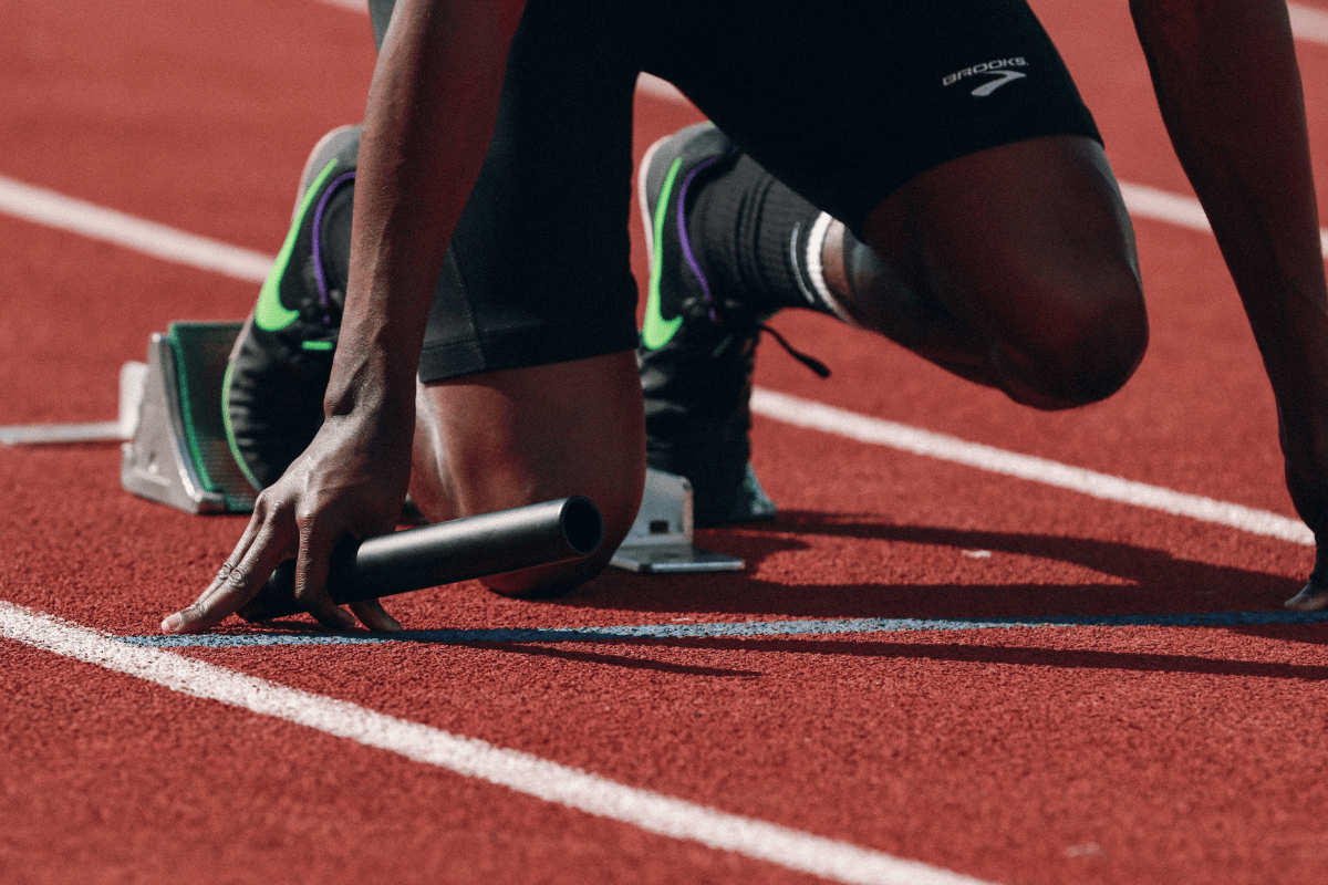 Close-up of a sprinter in the starting blocks holding a relay baton, showing the hip extension and anterior thigh loading position associated with quad dominance in track athletes.