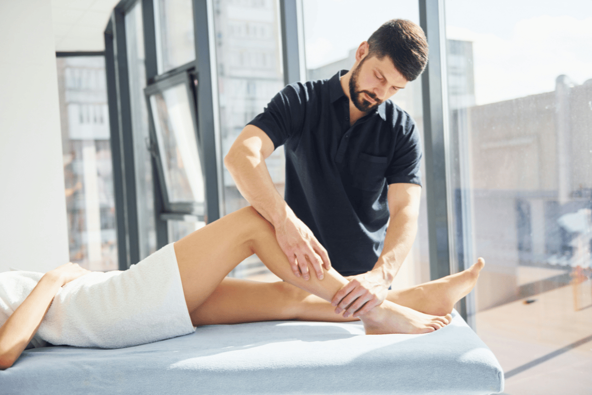Sports medicine clinician using both hands to manually assess a patient's lower leg and ankle on a treatment table, evaluating tibial stress syndrome and anterior compartment involvement.