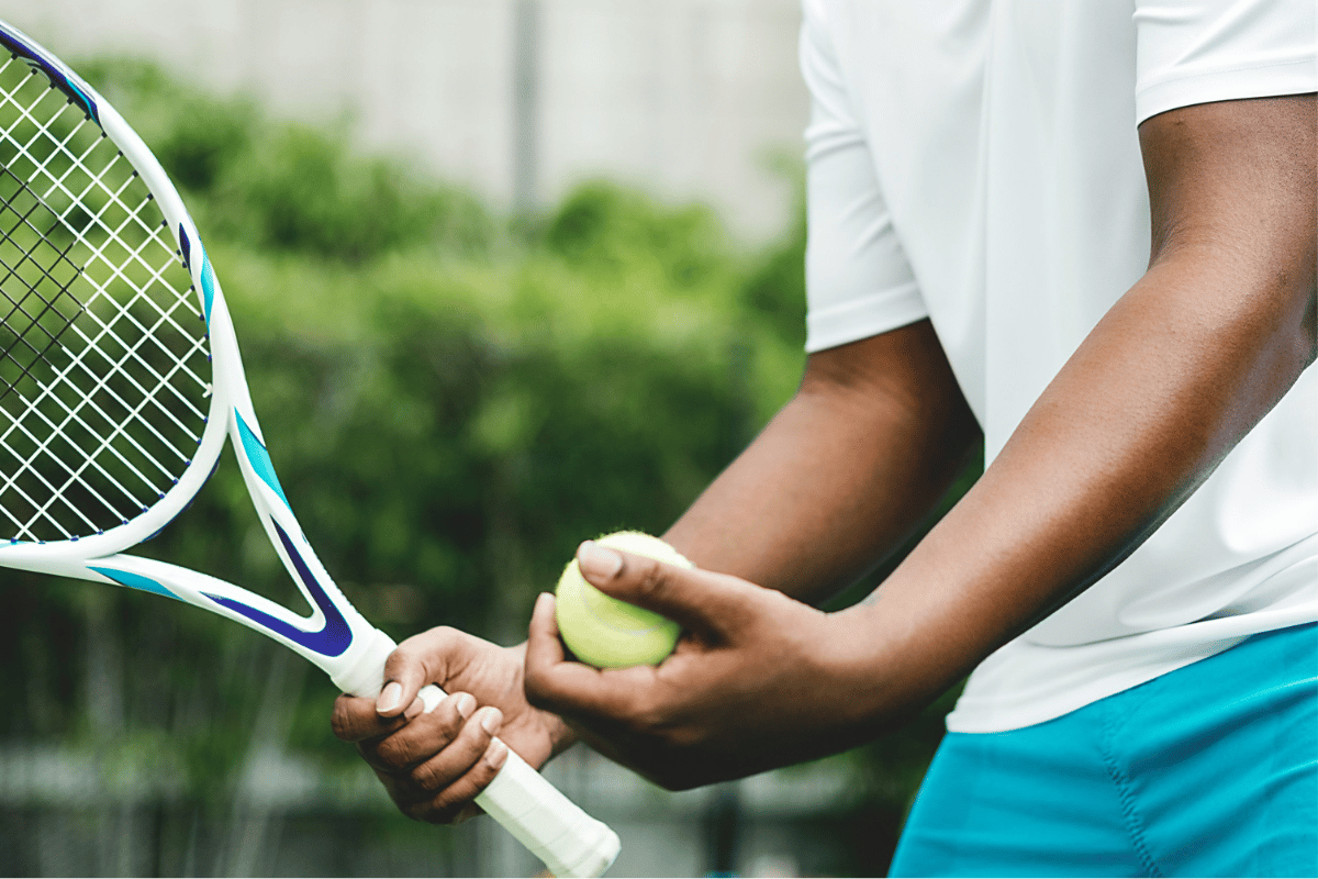 Tennis player gripping a racket handle and ball at the baseline, forearm engaged, illustrating grip-heavy athletic load