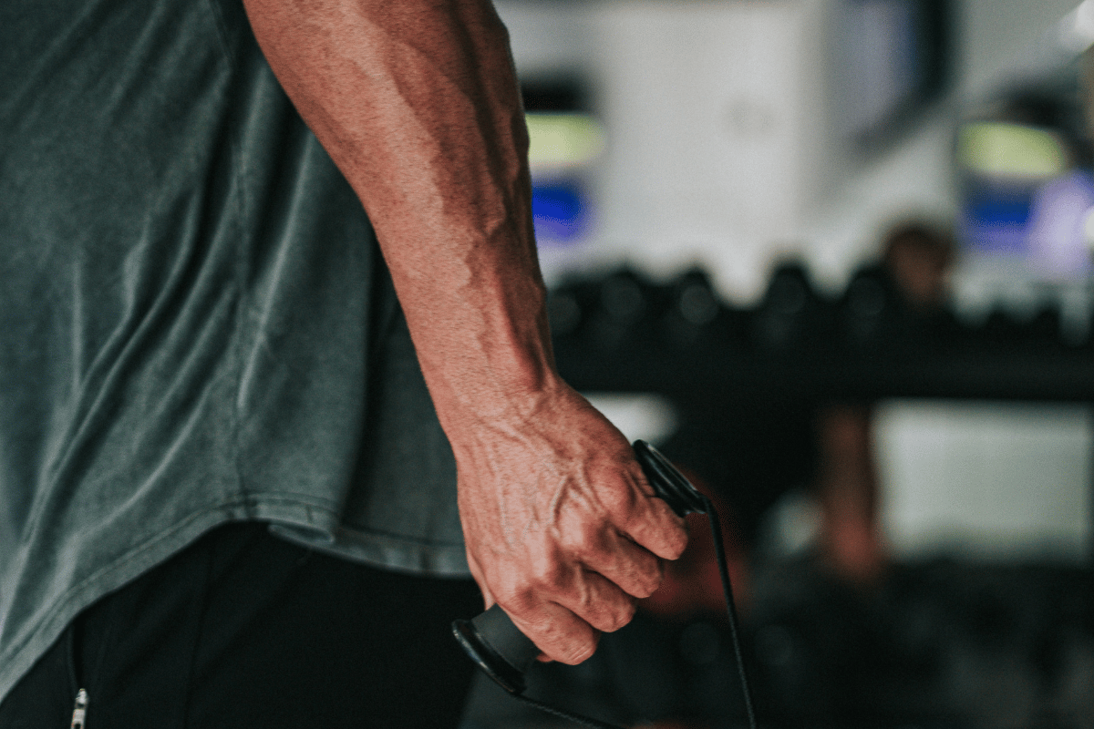 Close-up of an athlete's forearm and hand gripping a cable handle in a gym, flexor muscles and tendons visible, illustrating repetitive forearm grip load