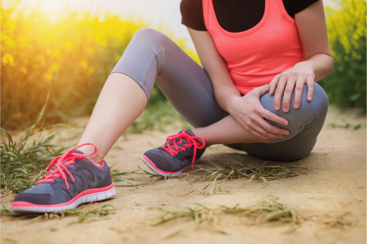 Runner pausing on a trail surrounded by wildflowers, gripping her knee and upper shin, illustrating the lower leg discomfort behind shin splints and tibial stress patterns.