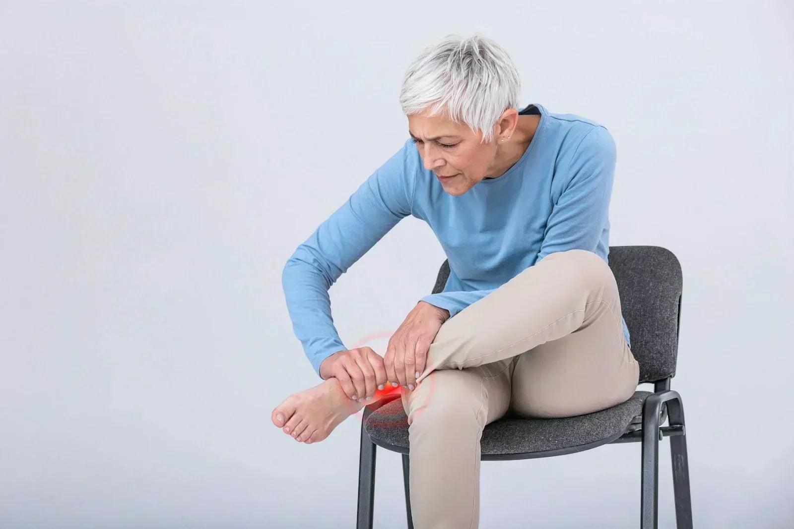 Older adult sitting in a chair holding her foot and ankle with visible discomfort, illustrating pain from a sprain or strain and the need for gentle recovery techniques.