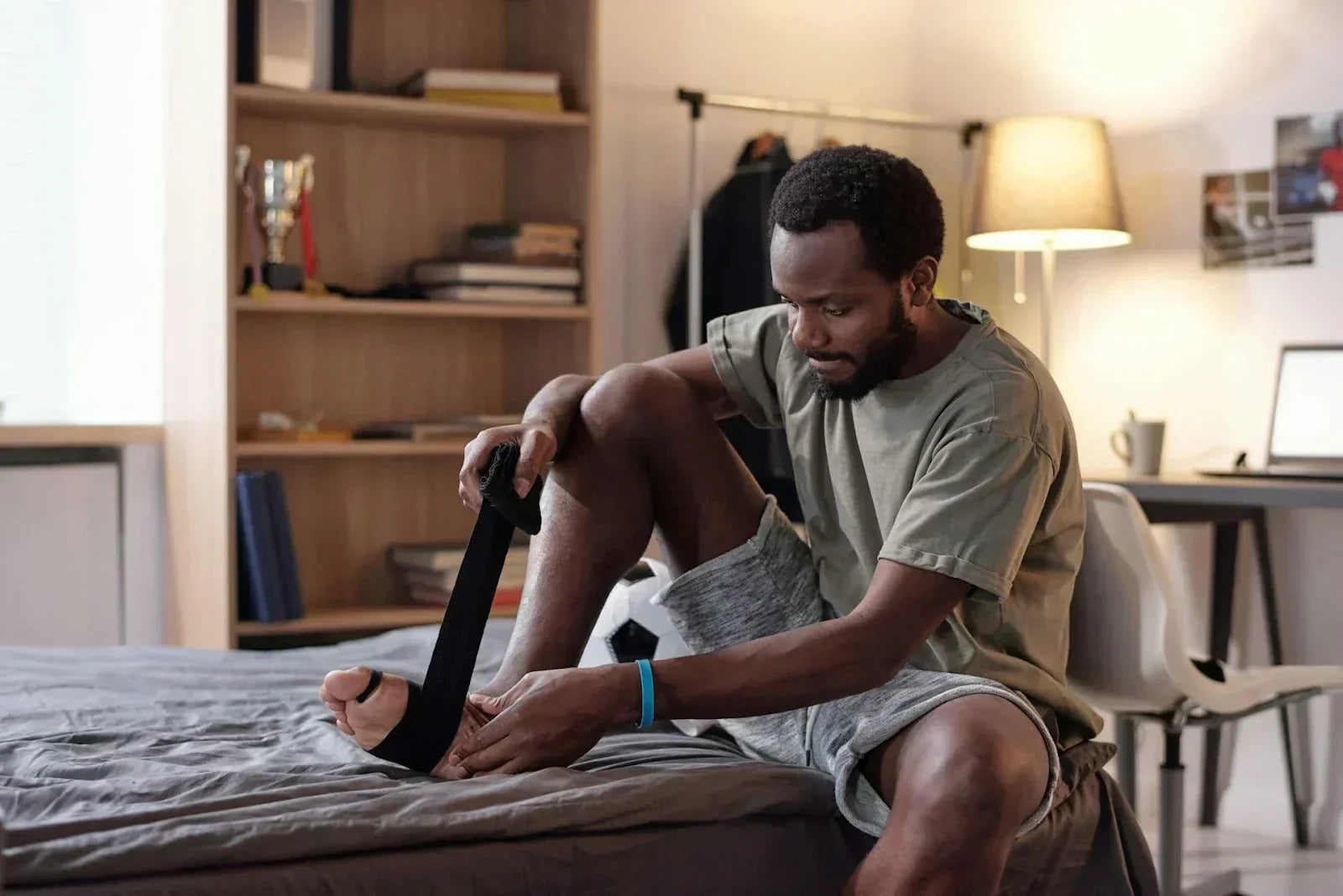 Male athlete sitting on a bed applying an ankle support strap, demonstrating at-home recovery for sprains and strains as part of a structured training and rehabilitation routine.